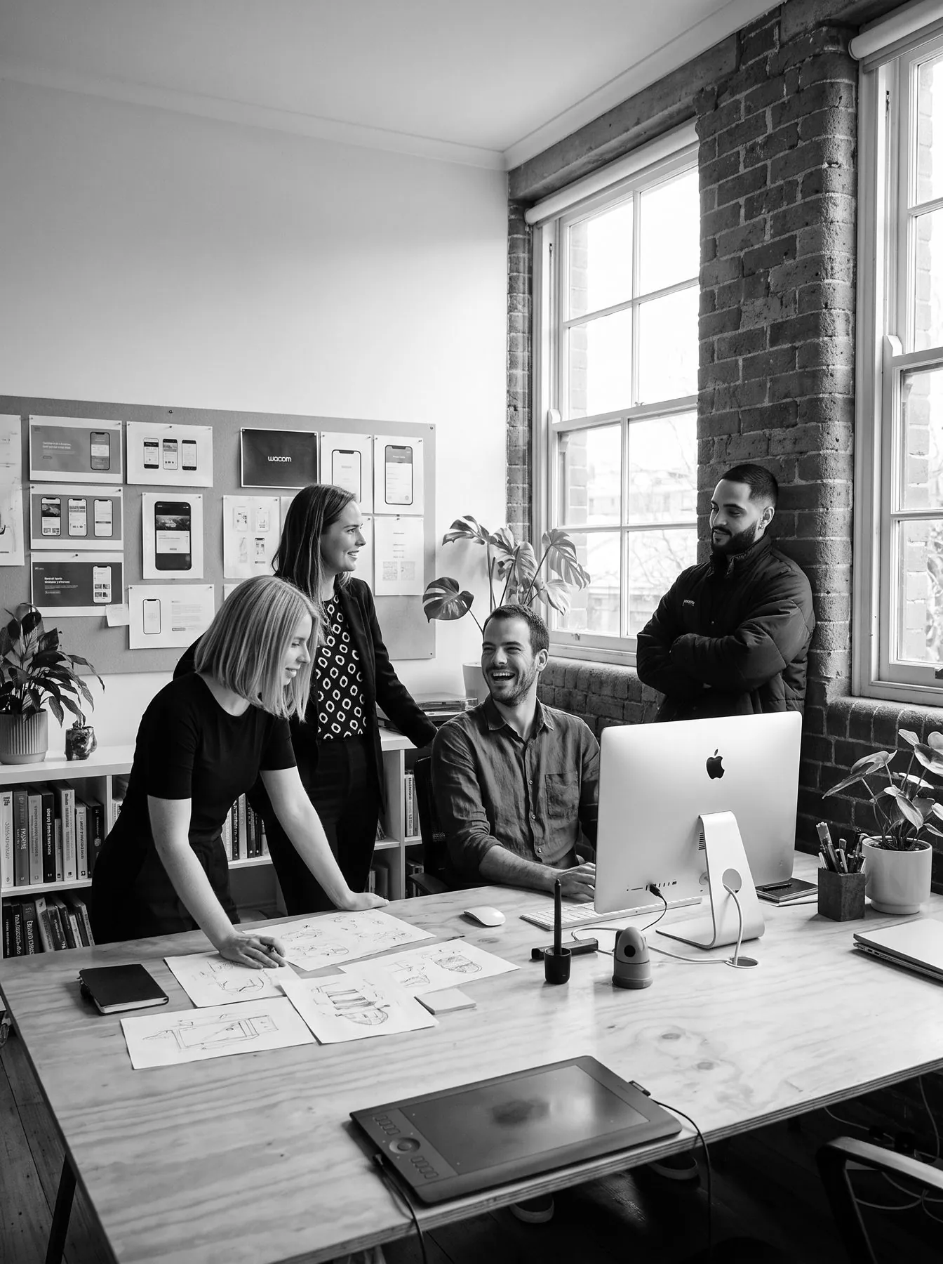 A design team laughing around a shared desk in a brick-walled studio